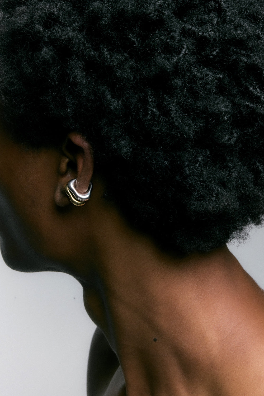 A close-up side profile of a person with dark curly hair wearing a chunky, multi-layered hoop earring in silver and gold-tone metals against a plain light gray background.