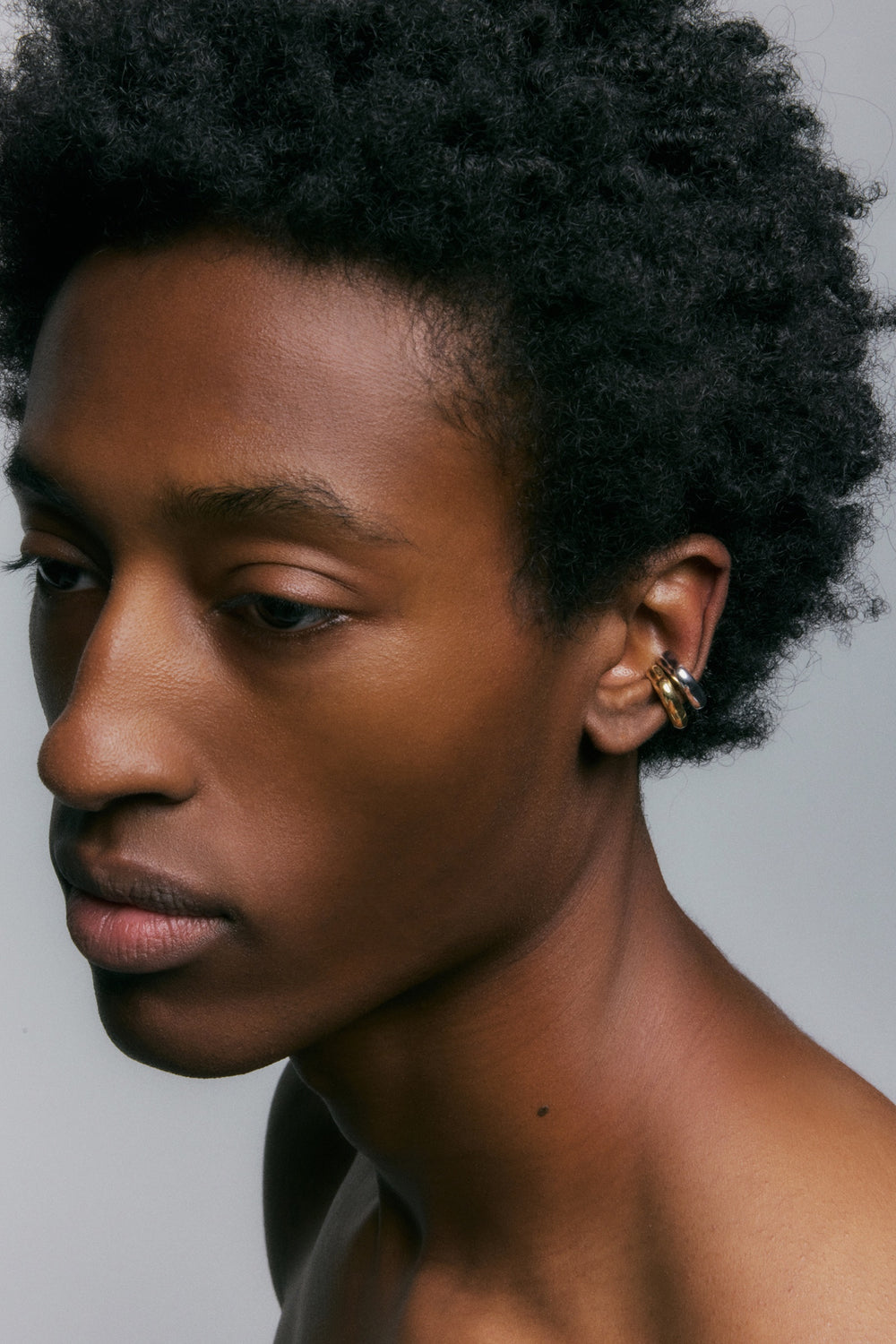 Close-up portrait of a person with dark curly hair wearing multiple gold hoop ear cuffs stacked on one ear, against a plain light gray background.